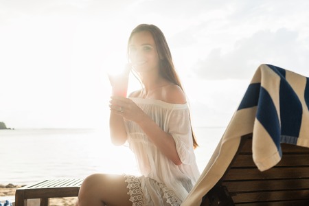 High-key portrait of a beautiful young woman smiling while enjoying a cocktail on a tropical beach during summer vacation in Flores Island, Indonesiaの写真素材