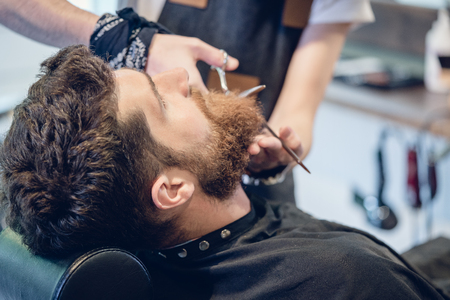 Close-up of the hand of a skilled barber using scissors, while trimming the beard of a young redhead customer in a trendy hair salon for menの写真素材