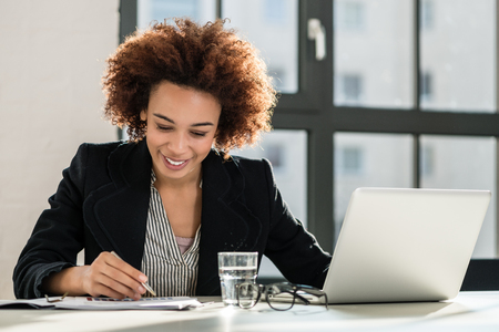 Portrait of African American female expert analyzing printed business report while sitting at desk in the officeの写真素材