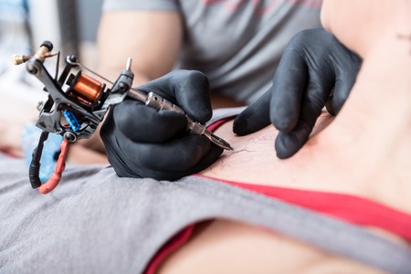 Close-up of the hands of a skilled artist using a modern machine, while making the contour of a cool tattoo on the chest of a female clientの写真素材