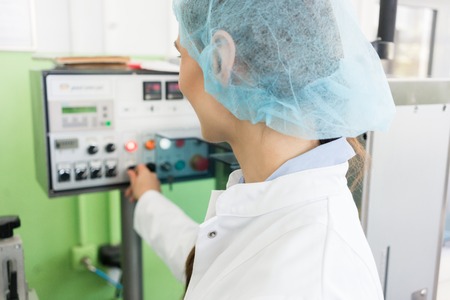 Experienced female manufacturing operator wearing protective headwear and white lab coat while controlling industrial equipment in a cosmetics factoryの写真素材