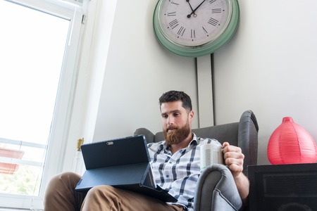 Young creative man sitting on armchair and holding a coffee mug while working on a tablet connected to internet in a modern co-working spaceの写真素材