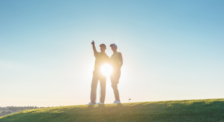 Low-angle view of the silhouette of a man pointing to the horizon, while standing next to his female partner on a professional golf course against sunshine and clear blue skyの写真素材