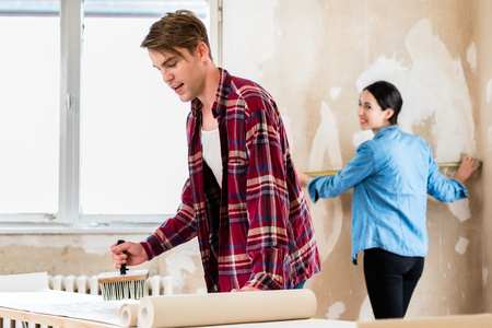 Young man applying adhesive to the back of the wallpaper sheet while his girlfriend is measuring the prepared wall in their new homeの写真素材