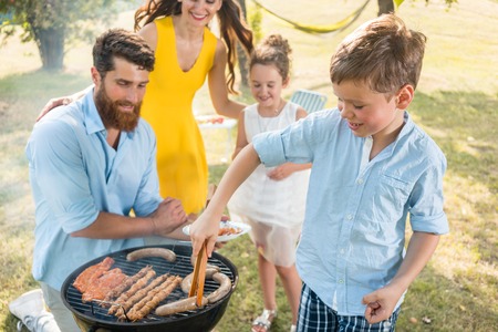 Father and son smiling while preparing meat on the charcoal barbecue grill during a relaxing family picnic outdoors in a summer dayの写真素材