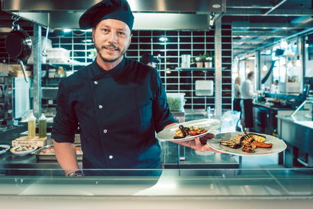 Portrait of a confident and experienced master chef holding two plates with tasty grilled vegetables behind the counter of a trendy restaurantの写真素材