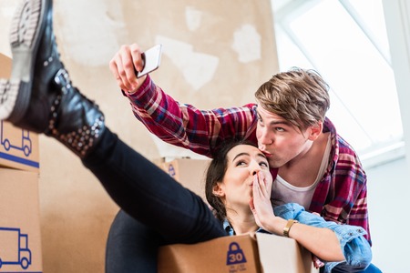 Happy young man and woman posing while making a selfie surrounded by boxes after moving in into a new homeの写真素材