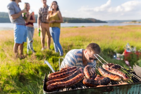 Barbecue party with grilled sausages at field near lake, men and womenの写真素材