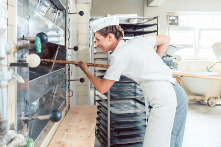 Baker woman getting bread out of bakery oven in her bakeryの写真素材