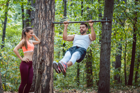 Woman watches man doing exercises on high bars in forest gym