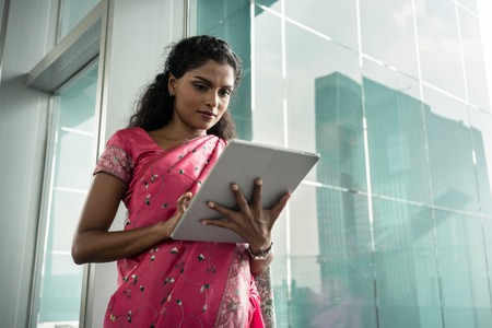 Portrait of young Indian woman using a tablet PC outdoors against the glass wall of a modern business buildingの写真素材