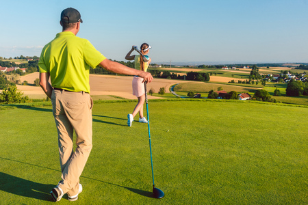 Full length rear view of a man wearing modern golf outfits, while watching his partner striking the ball during match on the green grass of a professional golf course in summerの写真素材