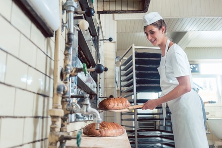 Baker woman getting bread out of bakery oven in her bakeryの写真素材