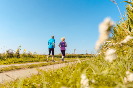 Full length rear view of two healthy senior people jogging on a country road against clear blue sky in summerの写真素材