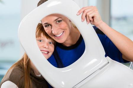 Funny portrait of a cheerful woman and her cute daughter smiling and looking at camera through a white toilet seat in a sanitary ware shopの写真素材
