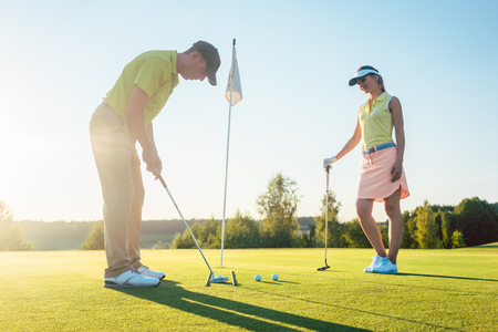 Full length side view of a man ready to hit the golf ball into the hole, while exercising the short shot with his game partner in a modern country club in summerの写真素材