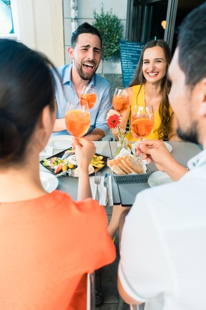 Two happy young couples with a trendy lifestyle toasting while sitting together at restaurant for a delicious lunch outdoors in summerの写真素材
