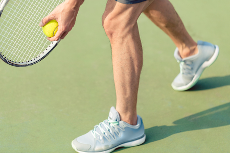 Low section of a professional player wearing gray sport shoes while holding the ball and the tennis racket during match on green surfaceの写真素材