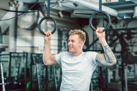 Portrait of a blond handsome young man with strong muscular arms smiling while hanging on gymnastic...