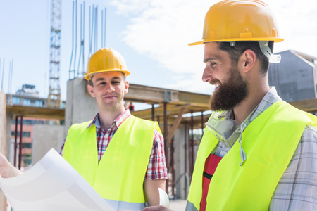 Two young construction workers wearing yellow hard hats and reflective safety vests while analyzing...