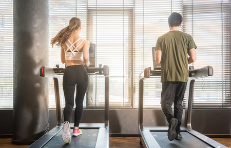 Full length rear view of young man and woman running on treadmills during workout session at the gymの写真素材