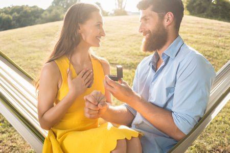 Side low-angle view of a cheerful young man making a marriage proposal to his beautiful girlfriend, while swinging together in a hammock outdoors in a summer dayの写真素材