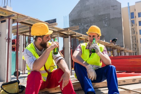 Two young workers smiling while drinking a cold alcoholic or non-alcoholic beer, during break at work on the construction site in a sunny day of summerの写真素材