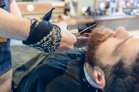 Close-up of the hand of a skilled barber using scissors, while trimming the beard of a young redhead customer in a trendy hair salon for menの写真素材