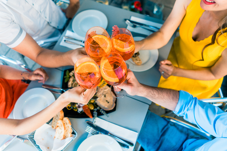 High-angle view of the hands of four friends holding stemmed wine glasses with a delicious orange summer drink, while toasting during lunch outdoors at a trendy restaurantの写真素材