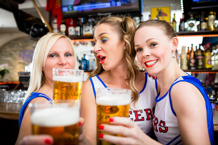 Cheerleader girls watching game of their favorite team in sport bar drinking beerの写真素材