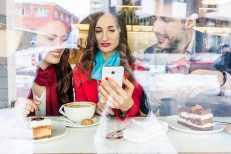 Fashionable young woman showing to her best friends pictures or videos on the mobile phone while sitting together in a trendy coffee shop downtownの写真素材