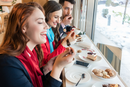 Side view of three young colleagues smiling happy while eating delicious cakes during break in a trendy coffee shopの写真素材