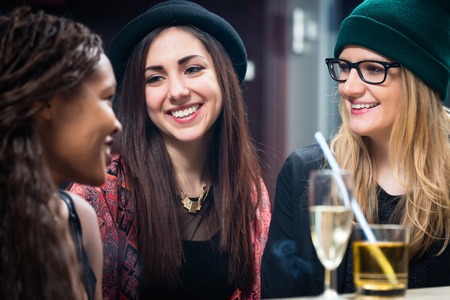 Three smiling female teenage friends looking at each other with glasses of drinks on table at...