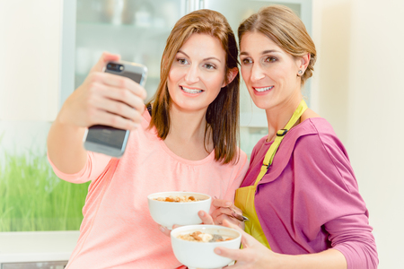 Two smiling young female friends holding bowl of oatmeal taking selfie on smart phoneの写真素材