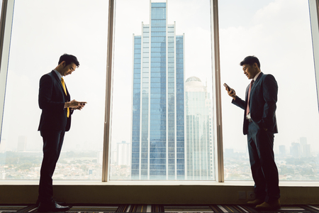 Side vie of two businessman using smart phone standing near the windowの写真素材