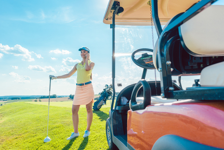 Full length of an active and cheerful woman, wearing modern golf apparel while talking on mobile phone on the golf course near the cart in a sunny day of summerの写真素材