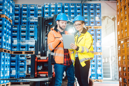 Workers in warehouse checking data on tablet computer with forklift in the background