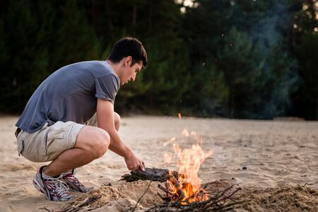 Young man preparing campfire on sand at beachの写真素材