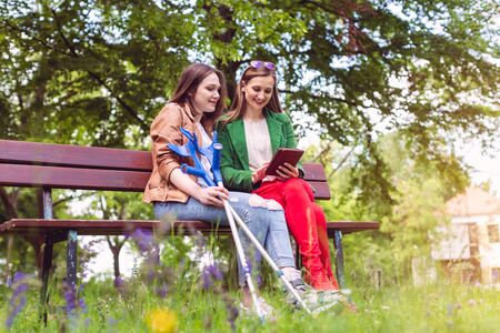 Two friends in a park, one with a broken feet and crutches reading a bookの写真素材