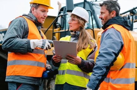 Three workers in a quarry discussing in front of heavy machinery looking at planの写真素材