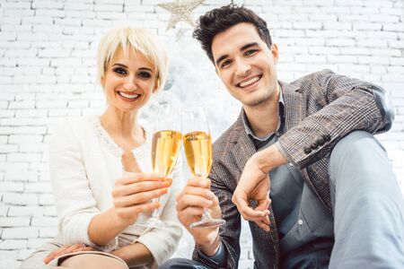 Couple clinking with sparkling wine for Christmas sitting in front of white treeの写真素材