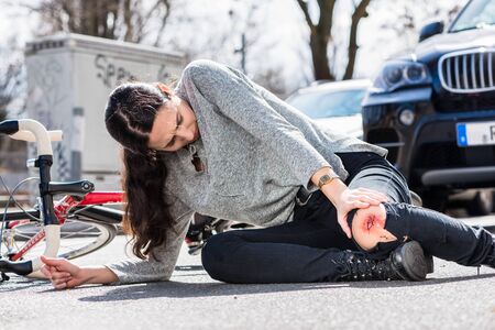 Young woman fallen down on the ground with a bleeding scrape on the knee after severe injury in bicycle accident on the streetの写真素材