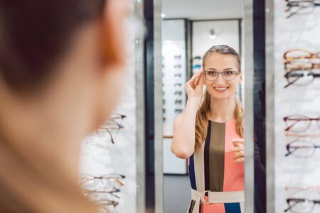 Young woman trying fashionable glasses in optometrist store looking at herself in the mirror