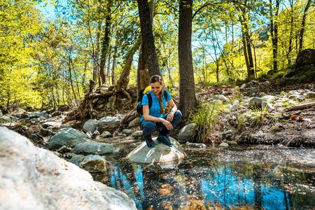 Hiking woman sitting on a stone in the middle of a creek passing the woods enjoying the spring