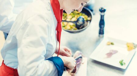 Chef cook in fancy restaurant kitchen slicing ham for a dish with plate of food in the backgroundの写真素材