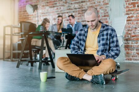 Hip worker in a startup coding with laptop sitting on skateboard with colleagues in backgroundの写真素材