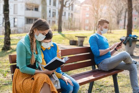 People sitting on park bench in the sun practicing social distancing in corona crisis wearing masksの写真素材