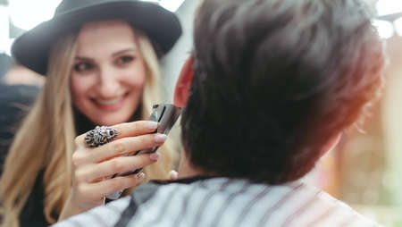 Barber woman trimming beard of client with the machine in her shopの写真素材