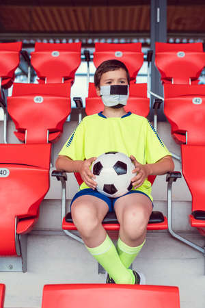 Lonely boy with football in his hand in soccer stadium watching a game wearing a maskの写真素材