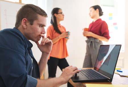 Young man working on laptop while his female colleagues standing with coffee cupの写真素材